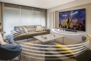 Imagem de uma sala de estar moderna com sofá e poltrona, uma mulher assistindo TV e ondas de som circulando o ambiente, representando imersão sonora, sem texto na cena.