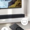 Soundbar preta posicionada abaixo de uma TV de tela plana em uma sala moderna, com controle remoto exibido em primeiro plano, mostrando conectividade e funções do aparelho.