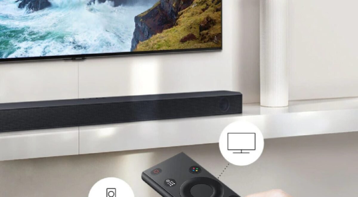 Soundbar preta posicionada abaixo de uma TV de tela plana em uma sala moderna, com controle remoto exibido em primeiro plano, mostrando conectividade e funções do aparelho.