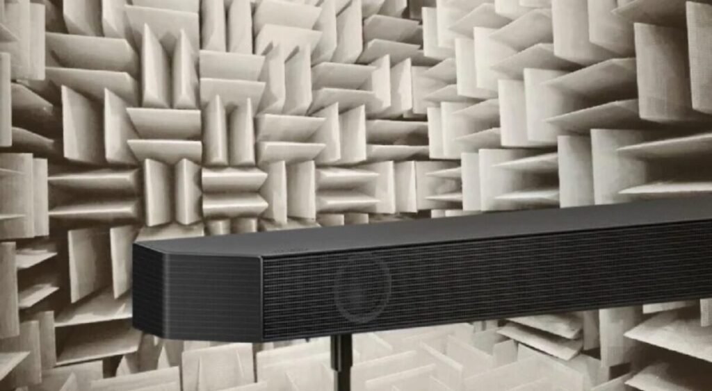 Soundbar preta posicionada em um suporte dentro de uma câmara anecoica com painéis acústicos brancos em forma de pirâmide ao fundo.